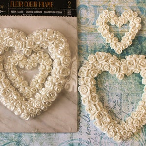 May include: Two white resin heart-shaped frames decorated with white roses. The frames are sold as a set of two. The larger frame is 7 inches wide and the smaller frame is 5 inches wide. The frames are labeled "Fleur Coeur Frame" and "Resin Frames, Cadres de Résine, Cuadros de Resina".