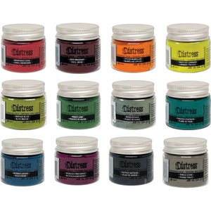Op de afbeelding: Twaalf kleine potjes Distress Oxide Stain, een soort verf, in verschillende kleuren. De potjes zijn gerangschikt in drie rijen van vier. Elk potje heeft een zilveren deksel en een wit etiket met het woord "Distress" en de naam van de kleur in het Engels en Frans.