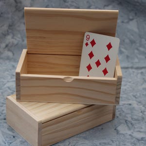 Może przedstawiać: Dwa niewykończone drewniane pudełka, jedno otwarte i jedno zamknięte, ułożone jedno na drugim. Górne pudełko zawiera kartę do gry, dziewiątkę karo. Pudełka są wykonane z jasnego drewna.