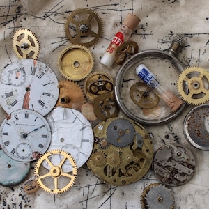 Puede incluir: Una colección de piezas de relojería y engranajes vintage, incluyendo una esfera redonda con números romanos, una esfera más pequeña con números arábigos y varios engranajes y piñones. Los objetos están dispuestos sobre un fondo de tela beige con marcas negras.