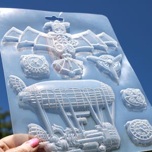 May include: A light blue, steampunk-themed mold with various designs, including an airship, clock faces, and a propeller. The mold is held up against a blue sky, showcasing the intricate details of the designs.