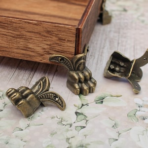 May include: Close-up of antique bronze furniture feet. The feet have an ornate, claw-like design. One is attached to a wooden box, while two others are separate. The background features a floral pattern. The feet are likely made of metal.