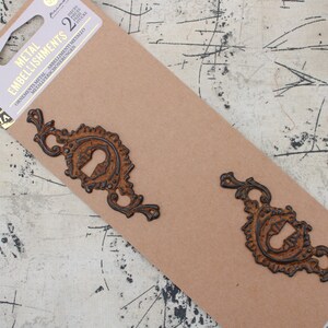 May include: Two ornate metal embellishments in a rusty brown color with black accents. The embellishments are packaged on a brown card with the text "Metal Embellishments" and "Ornamental Metal Abbellimenti Metallo Metallverzierung Schmuckverzierungen" printed on it.