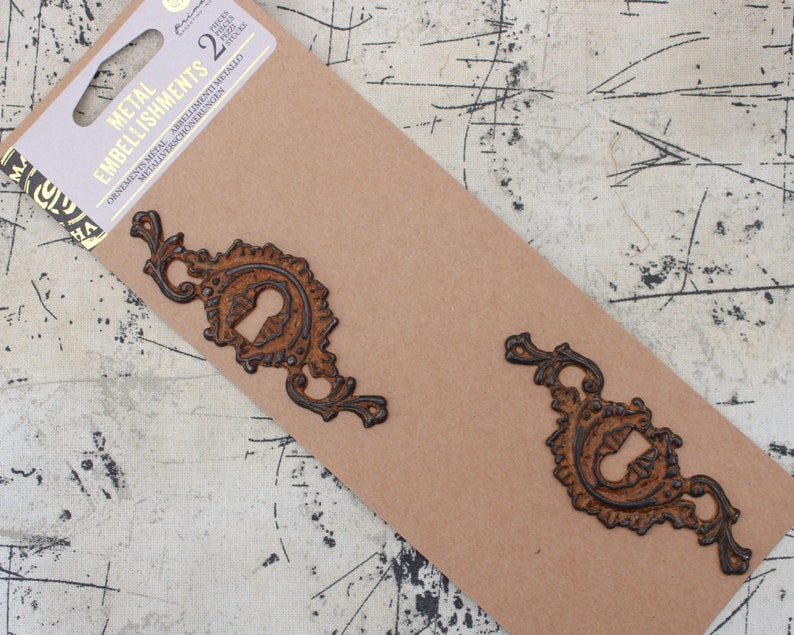 May include: Two ornate metal embellishments in a rusty brown color with black accents. The embellishments are packaged on a brown card with the text "Metal Embellishments" and "Ornamental Metal Abbellimenti Metallo Metallverzierung Schmuckverzierungen" printed on it.