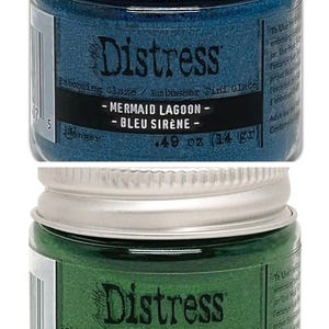 Op de afbeelding: Twee kleine potjes Distress Embossing Glaze. De bovenste pot is blauw en heeft het label "Mermaid Lagoon - Bleu Sir&egrave;ne - 0,49 oz (14 gr)". De onderste pot is groen en heeft het label "Mowed Lawn - Pousses de Gazon - 0,49 oz (14 gr)".