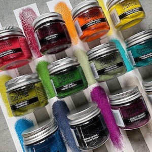 Op de afbeelding: Een verzameling van 10 kleine glazen potjes met zilveren deksels gevuld met kleurrijke glitter. Elk potje heeft een etiket met het woord "Distress" en een kleurnaam in het Engels en Frans. De kleuren omvatten Crushed Olive, Moved Lawn, Bungled Sage, Seedless Preserve, Hipped Sapphire, Spiced Marmalade, Aged Mahogany, Squeezed Lemonade, Peacock Feathers en Abandoned Coral.