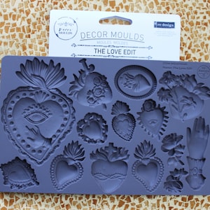 May include: A blue silicone mould featuring heart, hand, and floral designs. The mould is labelled "The Love Edit" and is for crafting. The background is a textured, golden surface.