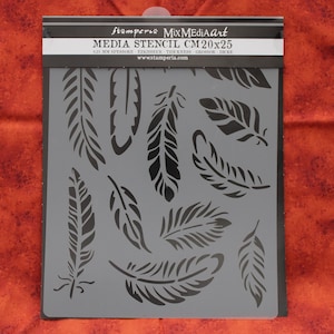 May include: A gray stencil with a feather design. The stencil is labeled "stamperia MixMediaArt MEDIA STENCIL CM 20x25". The feathers are black and vary in size and shape. The stencil is on a red-orange background.