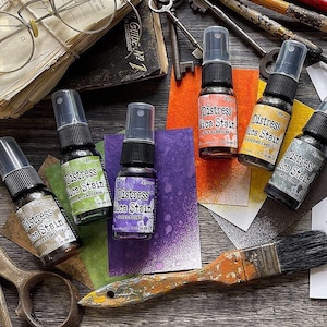 Op de afbeelding: Vier flesjes Distress Mica Stain in verschillende kleuren, waaronder paars, groen, oranje en zwart. Elk flesje heeft een spuitmond en een etiket met de productnaam en de kleur. De flesjes zijn op een houten oppervlak gerangschikt met andere knutselbenodigdheden, waaronder een kwast, sleutels en een boek.