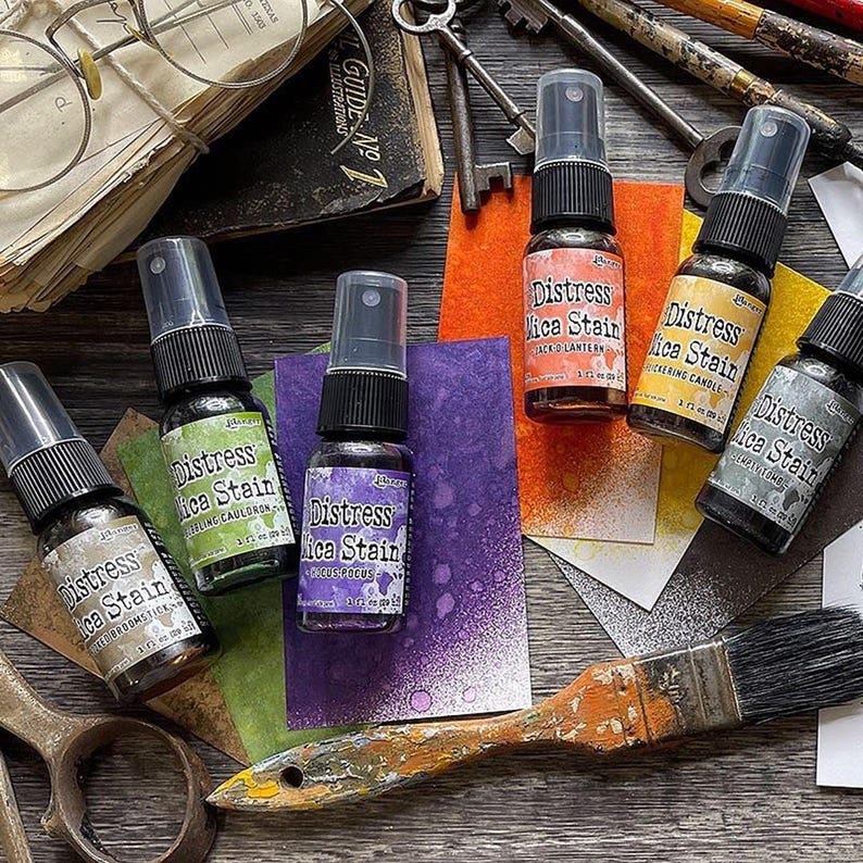 Op de afbeelding: Vier flesjes Distress Mica Stain in verschillende kleuren, waaronder paars, groen, oranje en zwart. Elk flesje heeft een spuitmond en een etiket met de productnaam en de kleur. De flesjes zijn op een houten oppervlak gerangschikt met andere knutselbenodigdheden, waaronder een kwast, sleutels en een boek.