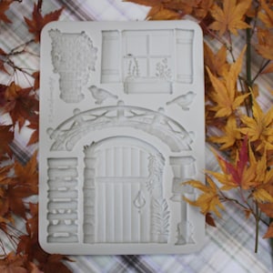 Może przedstawiać: Biała silikonowa forma z elementami architektonicznymi, takimi jak most, łukowate drzwi, okno i mur z cegieł. W projekcie uwzględniono również dwie małe figurki ptaków. Forma otoczona jest jesiennymi liśćmi.
