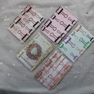 May include: Five sets of binder clips in different colours and finishes. Each set is arranged on a patterned card. The colours include black, gold, rose gold, green, and purple.