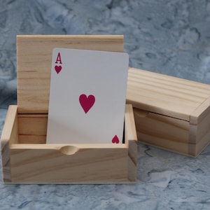 Może przedstawiać: Drewniane pudełko z przesuwaną pokrywą, obok otwartego pudełka z kartą do gry. Kartą jest As Kier, z dużym czerwonym sercem na środku i mniejszymi sercami. Pudełka wykonane są z jasnego drewna.
