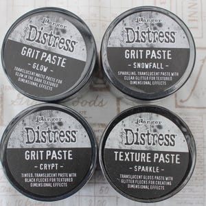May include: Four circular containers of Ranger Distress Grit Paste and Texture Paste. The containers are black with white text. The Grit Paste varieties include Glow, Snowfall, and Crypt. The Texture Paste is Sparkle.