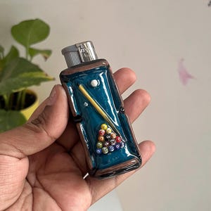 May include: A rectangular lighter featuring a three-dimensional design of a pool table with a teal felt surface, a yellow cue stick, and a triangular rack of colorful numbered billiard balls. The lighter has a metallic silver ignition top.