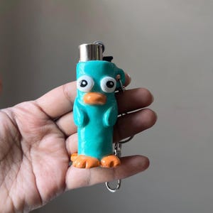 May include: A teal-coloured lighter case shaped like a cartoon platypus with large white eyes, black pupils, and an orange beak and feet. The cylindrical case features a metallic silver lighter top and is attached to a silver metal keychain ring.