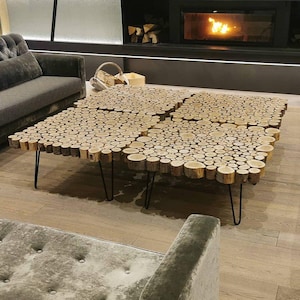 Puede incluir: Una mesa de centro única hecha de rodajas de madera natural dispuestas en un patrón de mosaico. La mesa tiene forma cuadrada con patas de horquilla de metal negro. La habitación cuenta con sofás grises y una chimenea.