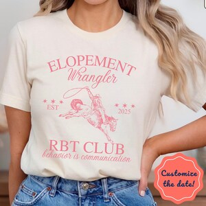 May include: A cream-coloured t-shirt with a pink graphic of a cowboy roping a horse, the text "ELOPEMENT Wrangler," and "RBT CLUB behavior is communication." The shirt also has the text "EST 2025."