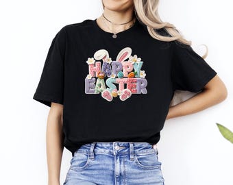 Może przedstawiać: Czarny t-shirt z kolorową grafiką "Happy Easter". Projekt zawiera uszy królika, jajka i kwiatowe akcenty. Tekst jest w stylu patchwork z różnymi wzorami i kolorami, idealny na obchody Wielkanocy.