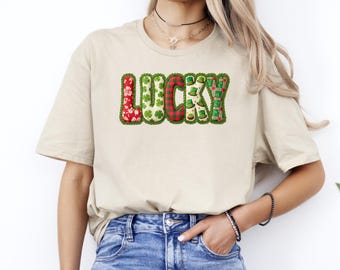 Może przedstawiać: Beżowa koszulka z napisem "LUCKY" w kolorowym patchworkowym wzorze. Litery zdobią koniczyny, wzory w kratę i zielone kapelusze krasnoludków, idealne na Dzień Świętego Patryka.