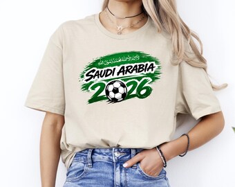 Może przedstawiać: Beżowa koszulka z zielono-białą grafiką. Grafika zawiera napisy "SAUDI ARABIA" i "2026" z piłką nożną pośrodku. Koszulka wykonana jest z miękkiego materiału.