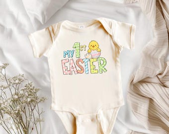 Może przedstawiać: Kremowe body dla dziecka z napisem "My 1st Easter" w kolorowych literach. Powyżej słowa "Easter" znajduje się rysunkowy kurczak wykluwający się z jajka. Body ma krótkie rękawy i jest wykonane z miękkiego materiału, idealne na święta Wielkanocy.