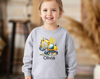Może przedstawiać: Jasnoszara bluza z żółtą koparką z uszami królika, zbierającą kolorowe pisanki. Imię "Olivia" jest wydrukowane poniżej. Bluza jest noszona przez małe dziecko.