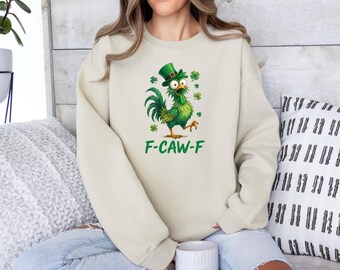 Może przedstawiać: Kremowa bluza z zielonym kogutem z kreskówki w kapeluszu krasnoludka i koniczyną. Tekst "F-CAW-F" jest wydrukowany na zielono pod kogutem. Bluza to swobodny, wygodny styl.