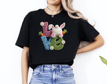 Może przedstawiać: Czarny t-shirt z kolorowym wzorem "LOVE" przedstawiającym dzierganego królika, marchewki i stokrotki. Litery są w różowy wzór w cętki, niebieskie kropki i zielony wzór w cętki. Uroczy t-shirt z grafiką w tematyce wielkanocnej.