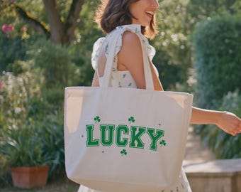 Może przedstawiać: Biała płócienna torba z napisem "LUCKY" w zielonych literach i zielonymi akcentami w postaci koniczyny. Torbę niesie osoba ubrana w białą sukienkę w kwiaty.