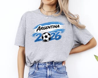 Może przedstawiać: Jasnoszara koszulka z napisem "Argentina" w kolorze czarnym, na niebiesko-białym wzorze pędzla. Poniżej znajduje się piłka nożna i liczba "2026" w kolorze niebieskim. Koszulka jest wykonana z miękkiego materiału.
