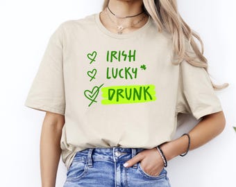 Może przedstawiać: Beżowa koszulka z zielonym napisem: "Irish, Lucky, Drunk." Słowo "Drunk" jest podświetlone w neonowo-zielonym prostokącie. Koszulka jest w stylu casual, odpowiednia na obchody Dnia Świętego Patryka.