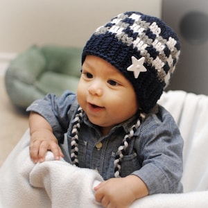 May include: A navy blue, gray, and white crocheted baby hat with a star embellishment. The hat has a checkered pattern and is worn by a baby wearing a denim shirt. The image includes the text "2014 Adorably Hooked".