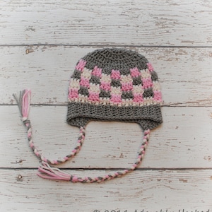 May include: A gray, pink, and white crochet hat with ear flaps and braided ties. The hat features a checkered pattern in pink and white, with a gray brim and crown. The braided ties end in tassels.