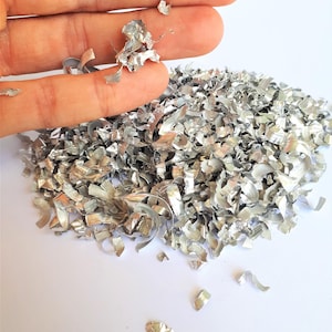 May include: Close-up of a pile of silver metal shavings, with a hand holding a small amount of the shavings. The shavings are curled and reflective, suggesting a metallic material. The background is a plain white surface.