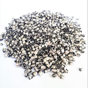 Op de afbeelding: Een close-up van een grote hoeveelheid kleine, glanzende metaalspanen. De spanen zijn zilverkleurig en hebben een cilindrische vorm met een gekruld, spiraalvormig uiteinde. Ze zijn verspreid over een wit oppervlak, waardoor een textuur en reflecterend uiterlijk ontstaat.