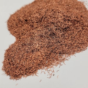 May include: A pile of copper-colored, fine, wire-like material. The material is densely packed, creating a textured, slightly uneven surface. The color is a warm, reddish-brown, and the overall appearance is of a loose, fibrous mass.