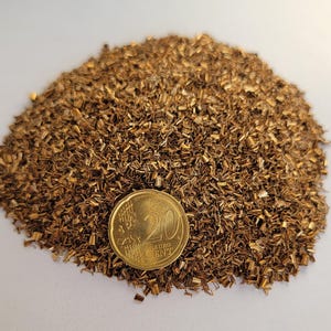 May include: A pile of small, irregular bronze-colored metal pieces. A gold-colored 20 Euro cent coin is placed on the pile for scale. The coin has the number 20 and the words "EURO CENT" on it. The background is a plain white surface.