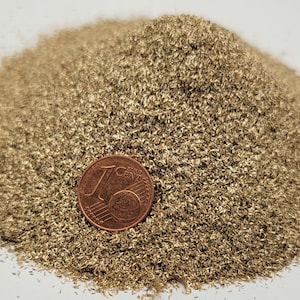 Può includere: Un mucchio di piccoli semi o granuli marrone chiaro, forse per l'artigianato o la decorazione. Una moneta da un centesimo di euro è posta di lato per il confronto delle dimensioni. I semi hanno un aspetto strutturato e granulare.