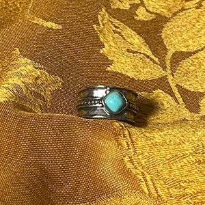May include: A silver ring with a turquoise stone. The ring has a detailed band with etched patterns and a central turquoise stone in a heart shape. The ring is set against a gold-colored, patterned background.