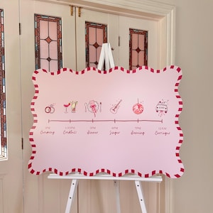 May include: A pink wedding schedule sign with a scalloped edge and red trim, displayed on a white easel. The sign shows illustrations and times for the ceremony, cocktails, dinner, singer, dancing, and carriages.