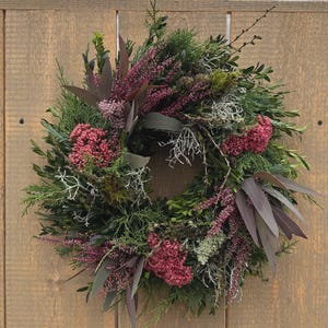 Könnte beinhalten: Ein kreisförmiger Kranz aus verschiedenen Grünpflanzen und Trockenblumen. Der Kranz weist eine Mischung aus grünem Laub, einschließlich immergrüner Zweige, und Akzenten aus bordeauxroten und rosa Blüten auf. Der Kranz wird vor einem hölzernen Hintergrund präsentiert.