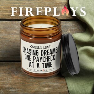 May include: A brown glass jar candle with a black lid. The candle has a white label with the text "Smells Like Chasing Dreams One Paycheck At A Time." The brand name "FIREPLOYS" is at the top. The candle is on a wooden surface.
