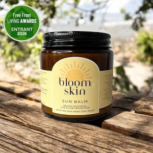 May include: A jar of Bloom Skin Sun Balm with a black lid and a yellow label featuring a sun graphic. The label states "Broad-Spectrum UVA & UVB Protection with 15% Non-Nano Zinc Oxide." A green "Free From Living Awards" badge is in the upper left.