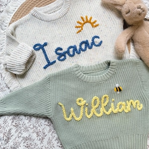 K&ouml;nnte beinhalten: Zwei Kinderpullover. Einer ist cremefarben mit dem Namen "Isaac" in Blau und einer gelben Sonne. Der andere ist salbeigr&uuml;n mit dem Namen "William" in Gelb und einer Biene. Ein Stoffhase ist im Hintergrund.