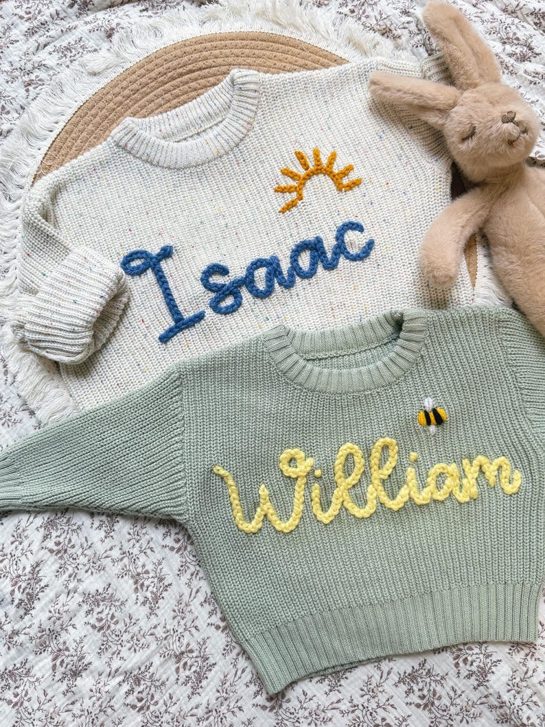 K&ouml;nnte beinhalten: Zwei Kinderpullover. Einer ist cremefarben mit dem Namen "Isaac" in Blau und einer gelben Sonne. Der andere ist salbeigr&uuml;n mit dem Namen "William" in Gelb und einer Biene. Ein Stoffhase ist im Hintergrund.