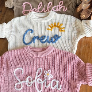 K&ouml;nnte beinhalten: Drei Strickpullover in den Farben Beige, Creme und Rosa. Jeder Pullover hat einen aufgestickten Namen auf der Vorderseite: "Delilah" in Rosa, "Crew" in Blau mit einer gelben Sonne und "Sofia" in Wei&szlig; mit einer kleinen Blume. Die Pullover werden auf einer Holzoberfl&auml;che pr&auml;sentiert.