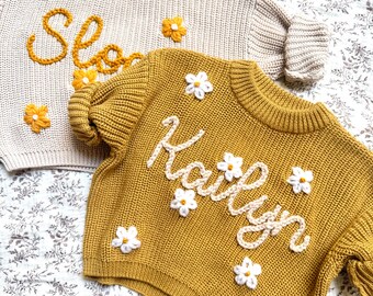 May include: Two children's sweaters. One is beige with the name "Slo" embroidered in yellow, and yellow flower accents. The other is mustard yellow with the name "Kaitlyn" embroidered in white, and white flower accents.