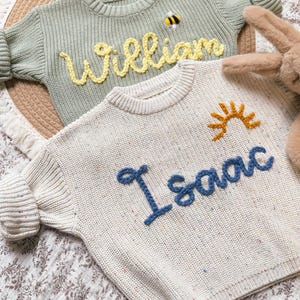 Könnte beinhalten: Zwei Kinderpullover. Einer ist hellgrün mit dem Namen "William" aus gelbem Garn und einer Bienenapplikation. Der andere ist cremefarben mit dem Namen "Isaac" aus blauem Garn und einer Sonnenapplikation.