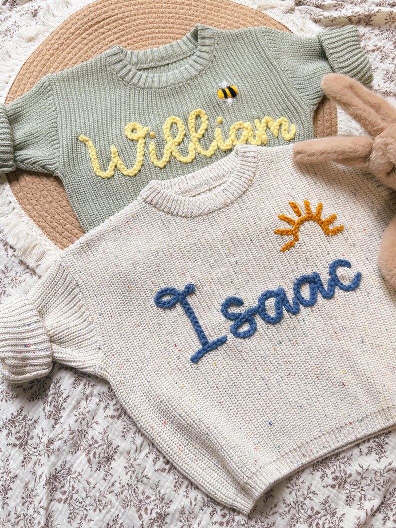 K&ouml;nnte beinhalten: Zwei Kinderpullover. Einer ist hellgr&uuml;n mit dem Namen "William" aus gelbem Garn und einer Bienenapplikation. Der andere ist cremefarben mit dem Namen "Isaac" aus blauem Garn und einer Sonnenapplikation.